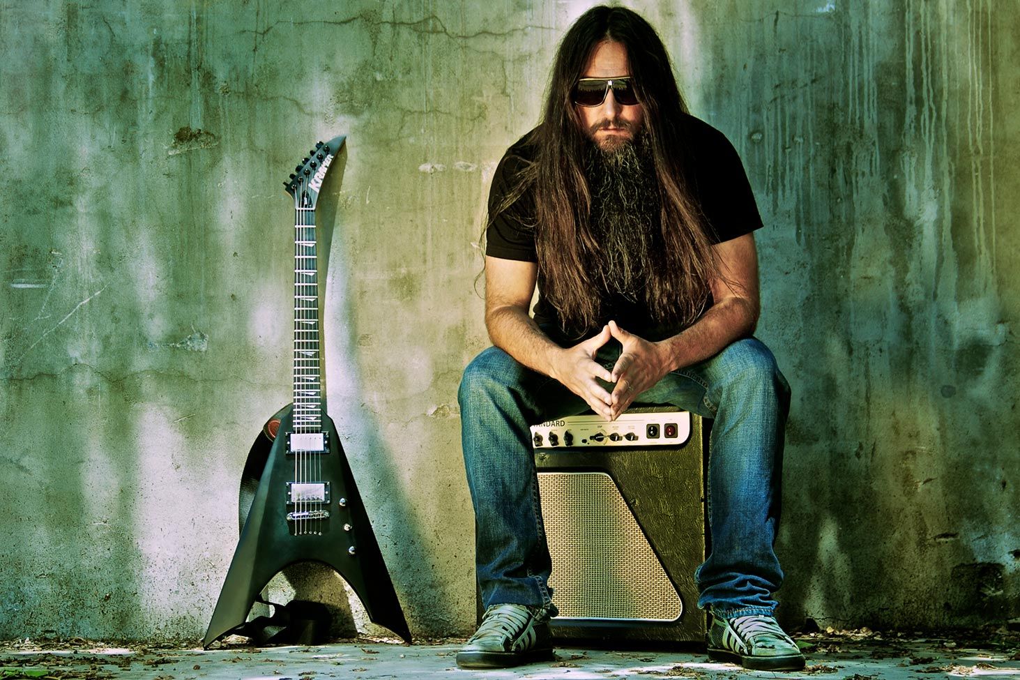 Rick Jacket musician portrait sitting on amplifier in front of green blue and yellow textured wall flying v guitar leaning against wall