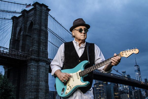Musician photo Robert Allen standing in front of Brooklyn Bridge wearing sunglasses and black hat while holding blue guitar at sunrise