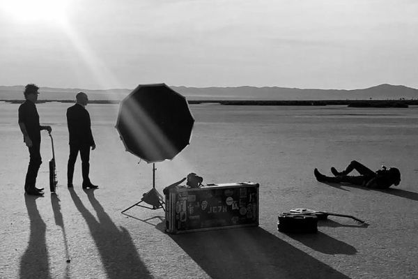 black and white bts photo Mark Maryanovich lying on ground while photographing 7Horse standing in desert guitar and case beside reflector