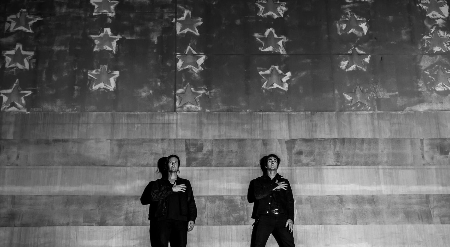 Band portrait Los Angeles Walking Papers both members standing in long shot against American Flag painted on wall hands across their hearts