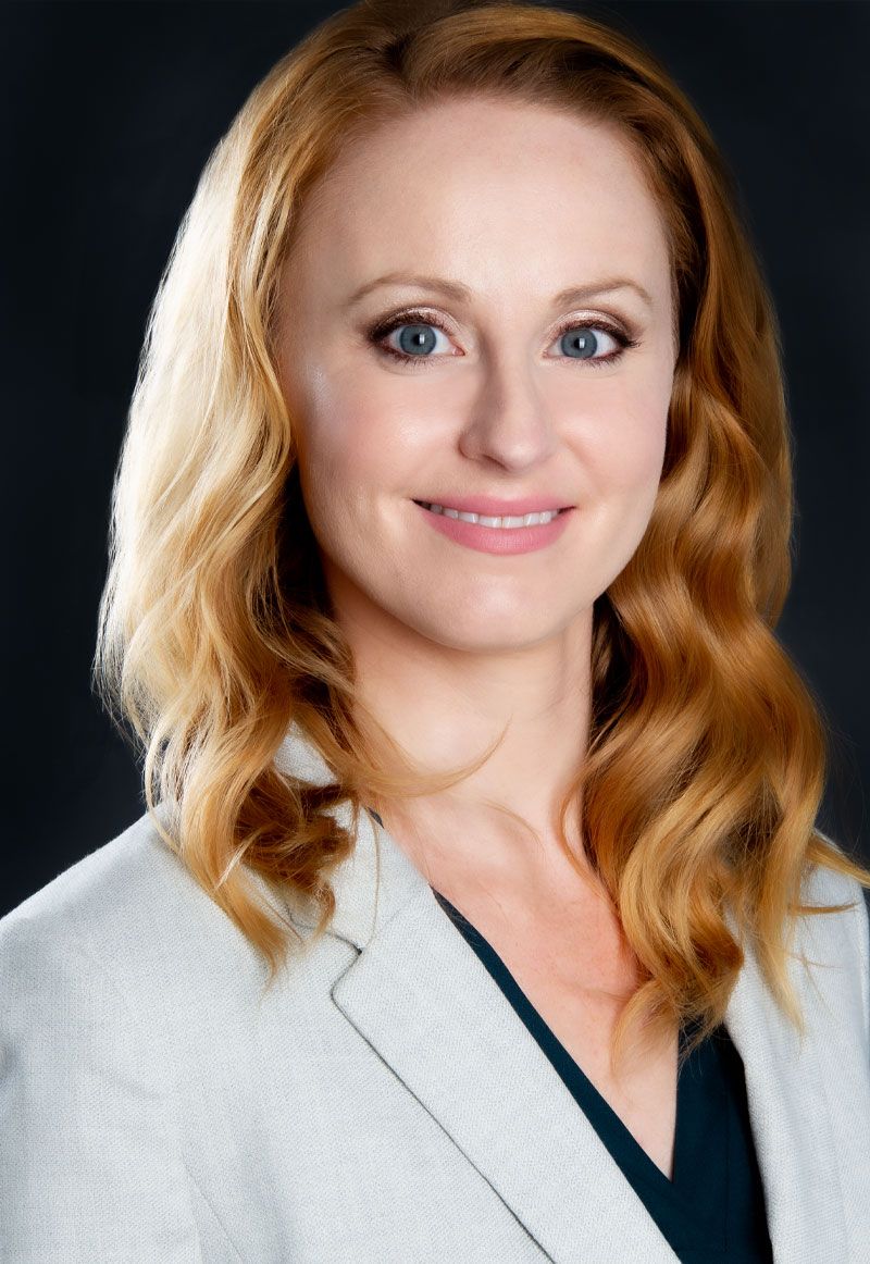 Professional headshot of a woman with wavy blonde hair wearing a light blazer against a dark background.