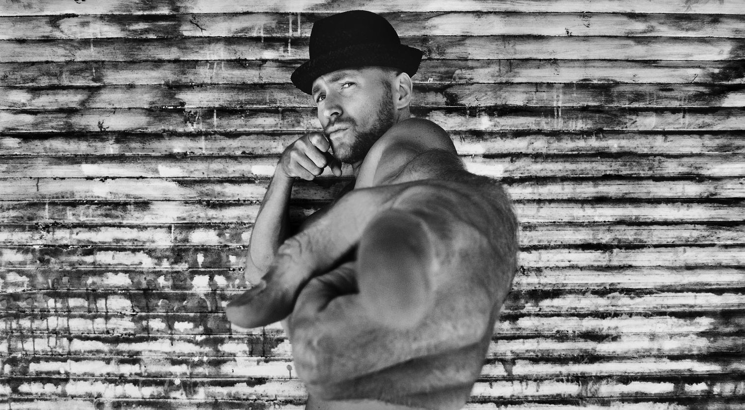 Male musician portrait Kyprios black and white pointing towards lens wearing fedora against dirty garage door