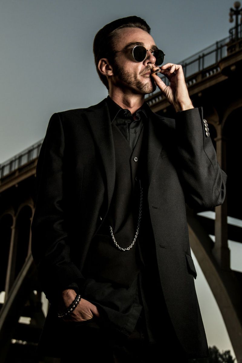 Professional Branding Portrait Author Chandler Morrison standing beneath bridge while smoking wearing black suit with pocket watch chain