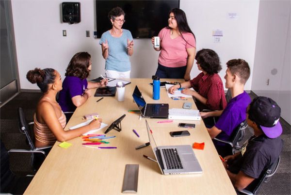 Smiling students sitting around table on campus listening to teacher