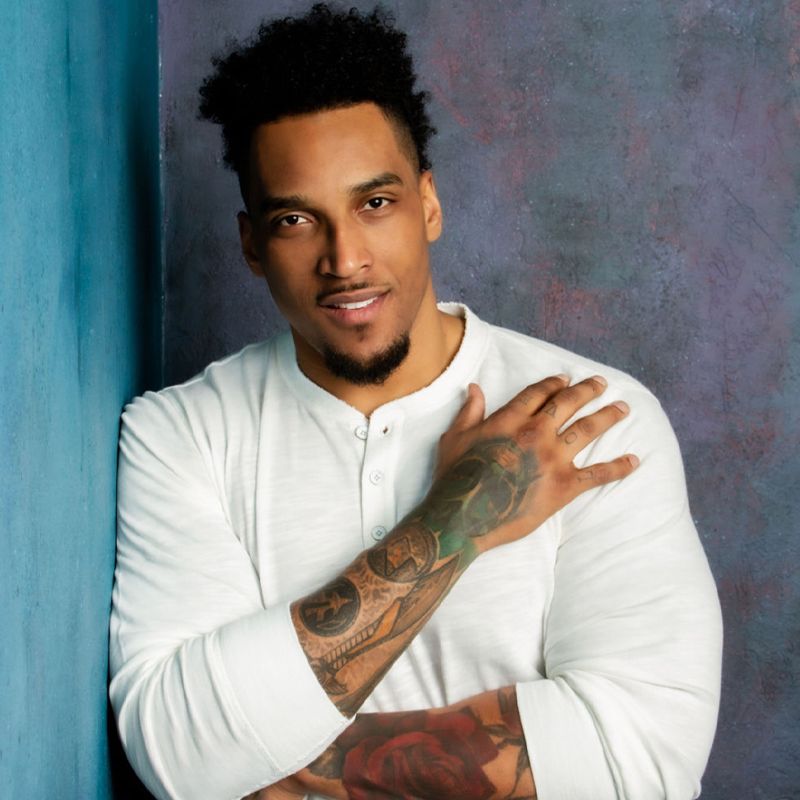 Personal branding headshot Joe Barksdale leaning against light blue wall wearing white long sleeve shirt smiling