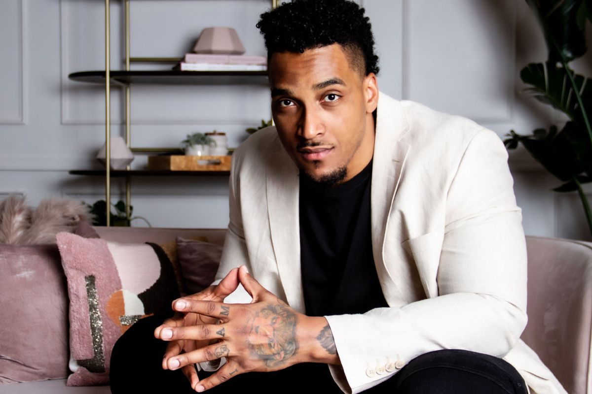 Branding portrait Joe Barksdale sitting on couch fingertips together weaing off white blazer black shirt and black pants