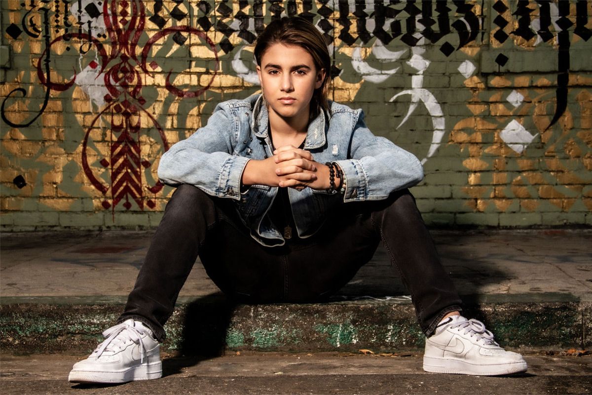 Preteen personal branding photo young boy sitting on curb hands clasped resting on knees graffiti wall behind him