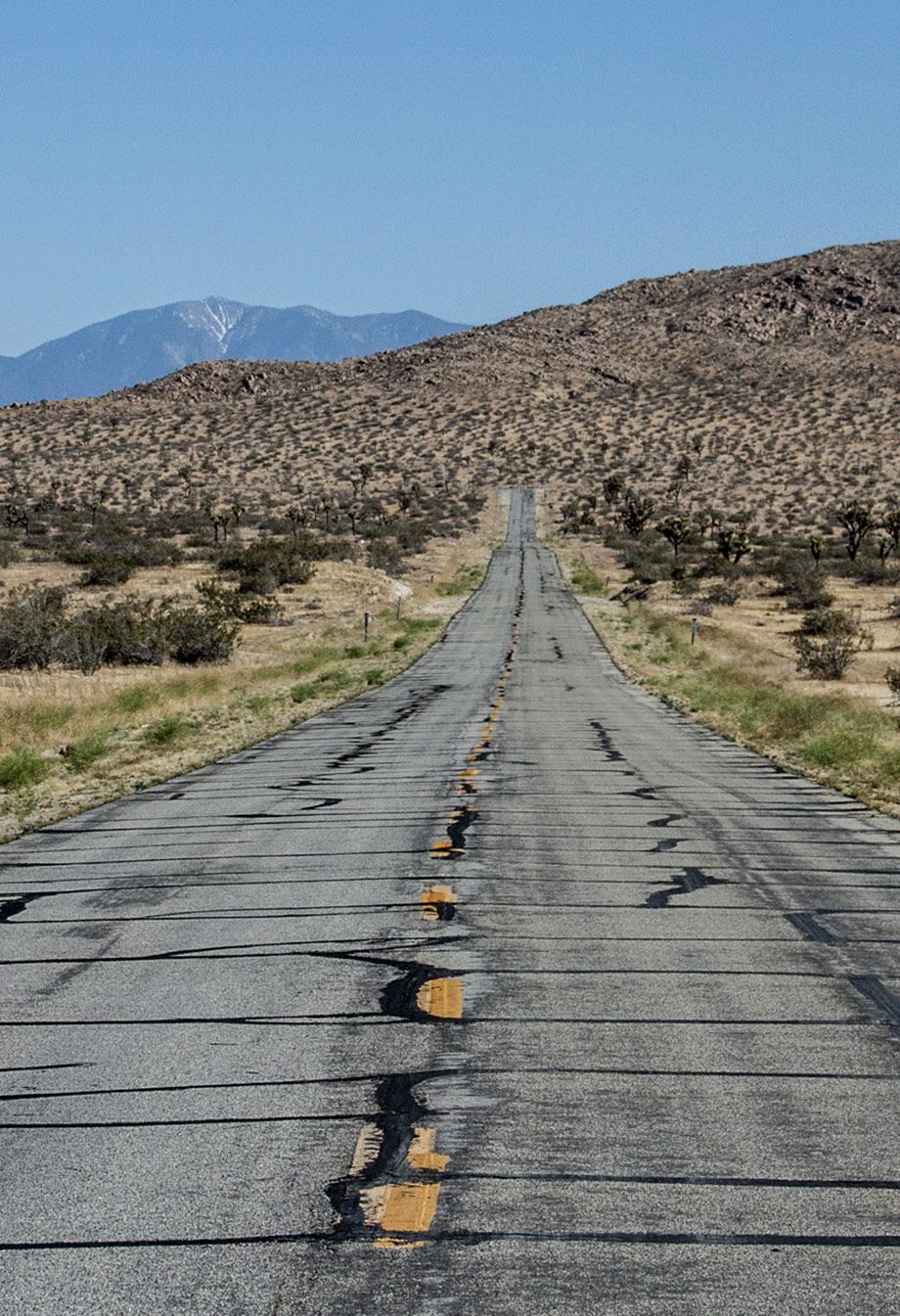 Long desert highway stretching toward mountains with cracked pavement and yellow lines.