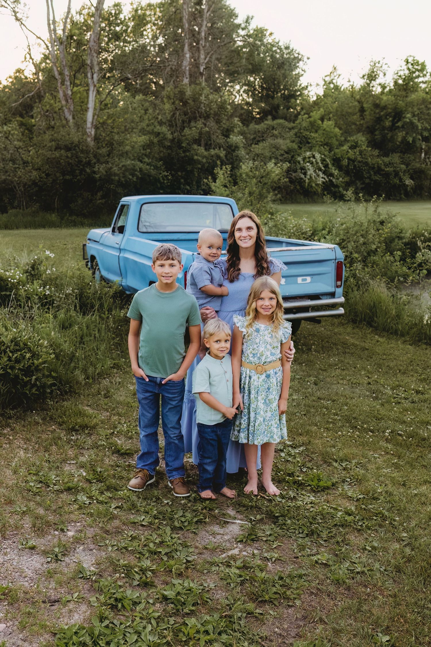 A mother poses with four children in front of a vintage blue pickup truck in a lush green field at sunset.
