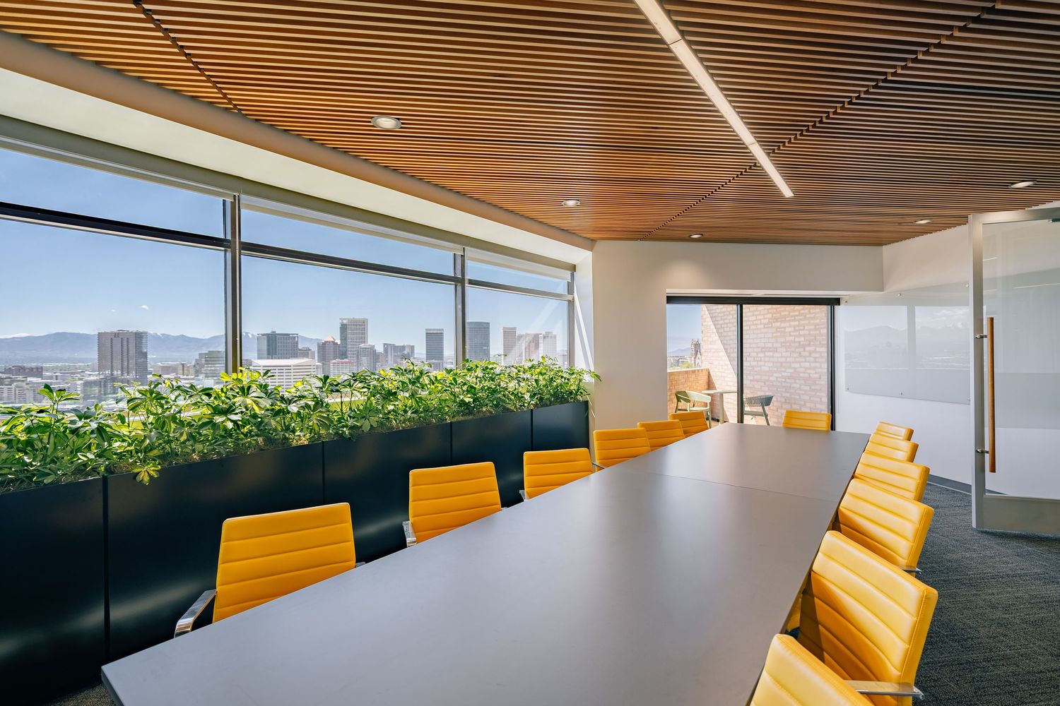 Modern conference room with yellow chairs, long table, green plants, wood ceiling, and city skyline views through floor-to-ceiling windows.