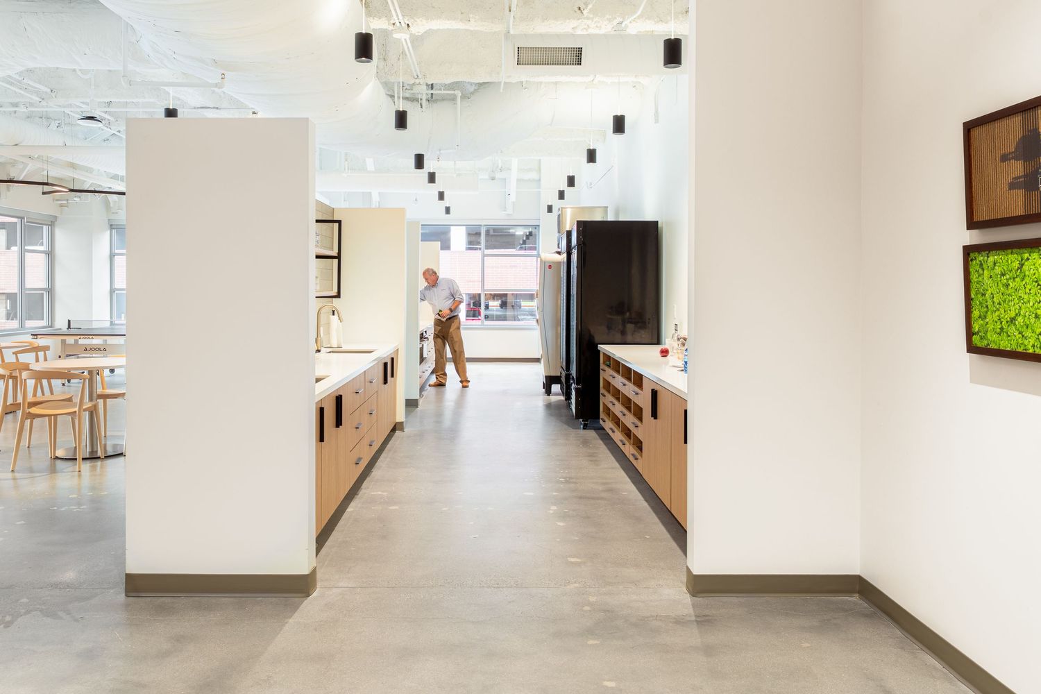 Modern office hallway with light wood cabinets, white walls, and pendant lighting leading to an open workspace area.