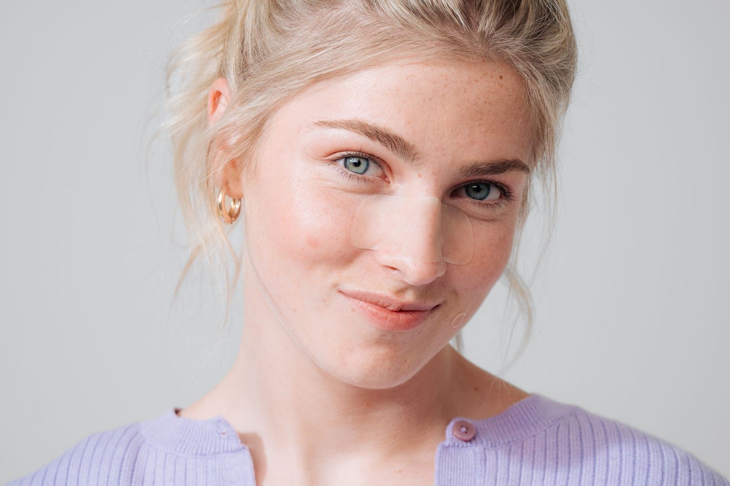 Close-up portrait of someone wearing a lavender sweater and gold earrings against a light gray background.