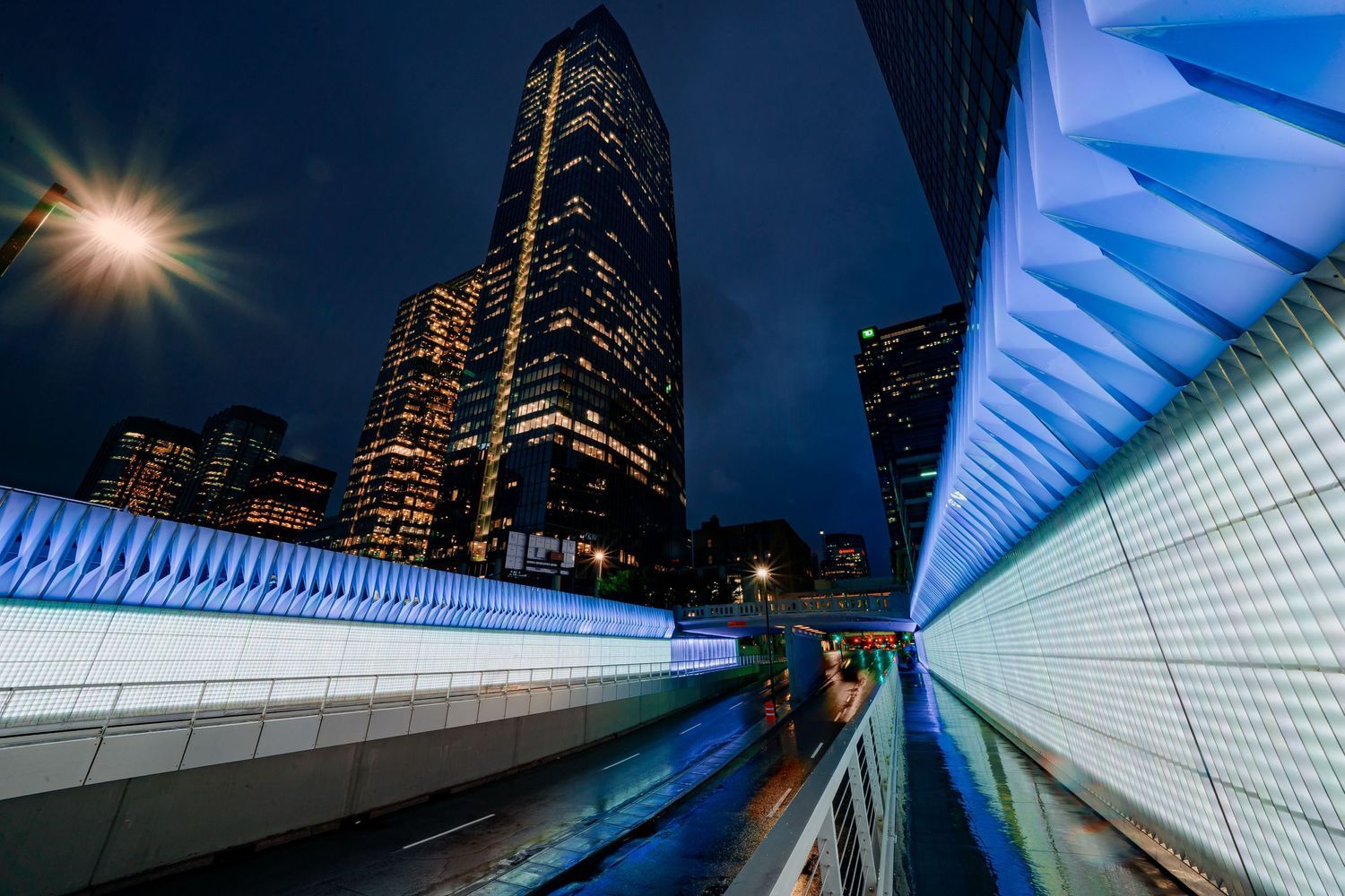 Modern architectural tunnel with illuminated white walls and blue lighting leads to city skyscrapers at night.