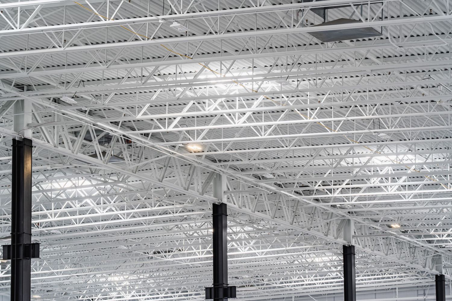 Industrial warehouse ceiling with exposed steel trusses, beams, and metal framework creating geometric patterns overhead.