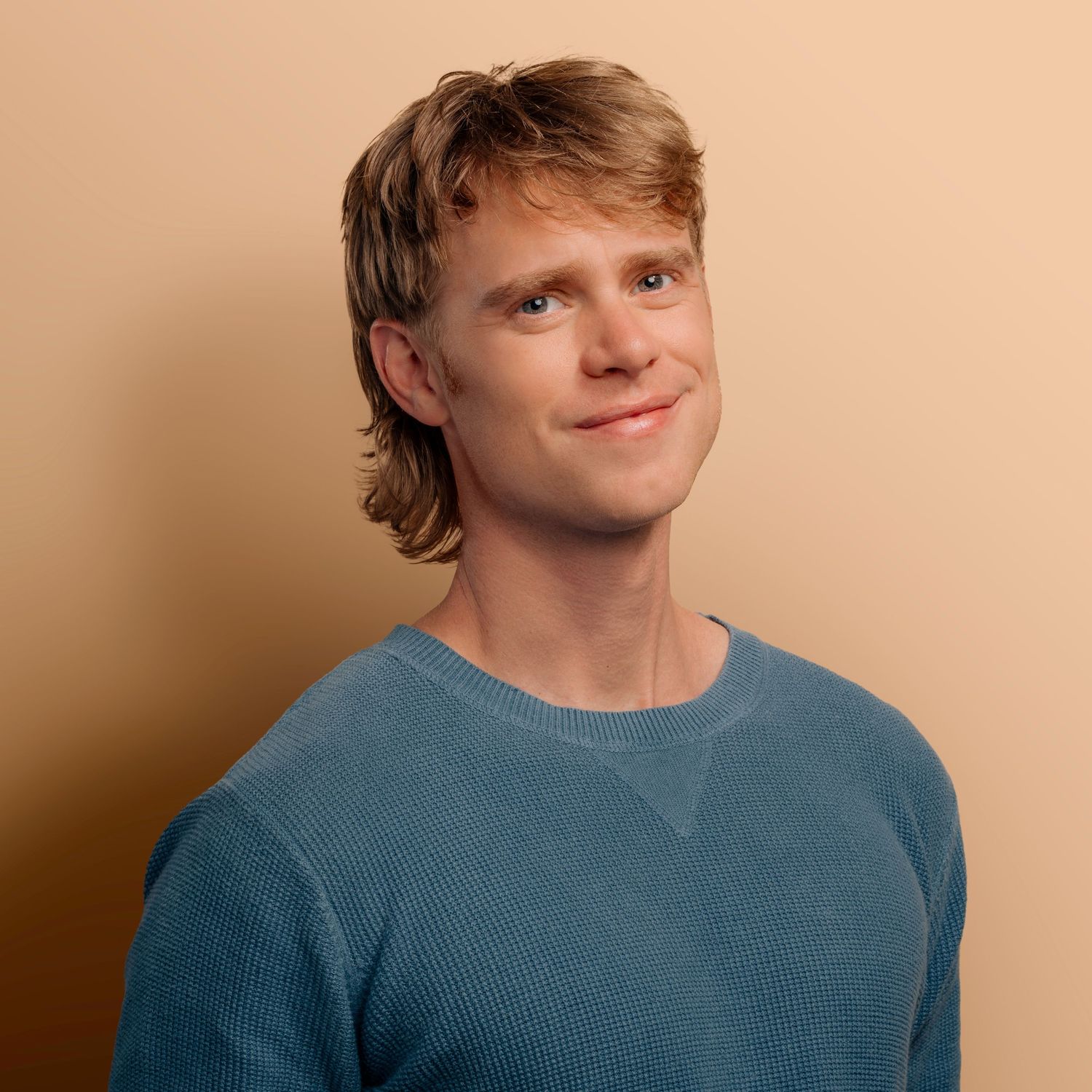 Portrait of a young man in blue sweater smiling against beige background.