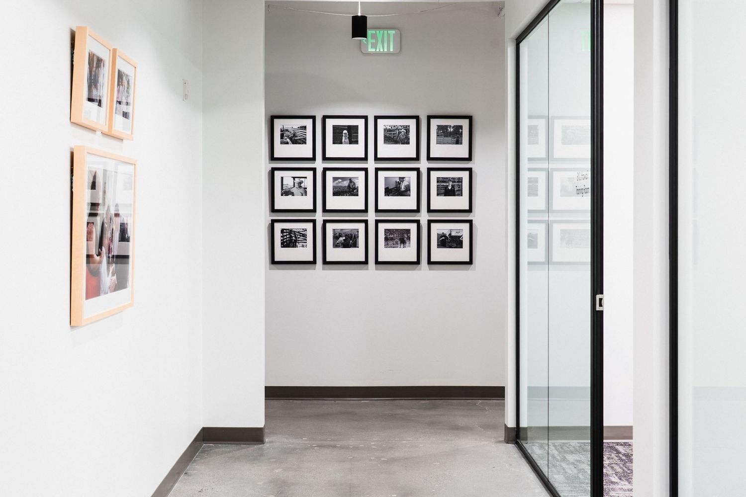 Modern office hallway with white walls featuring a grid of black and white framed photographs and glass doors.