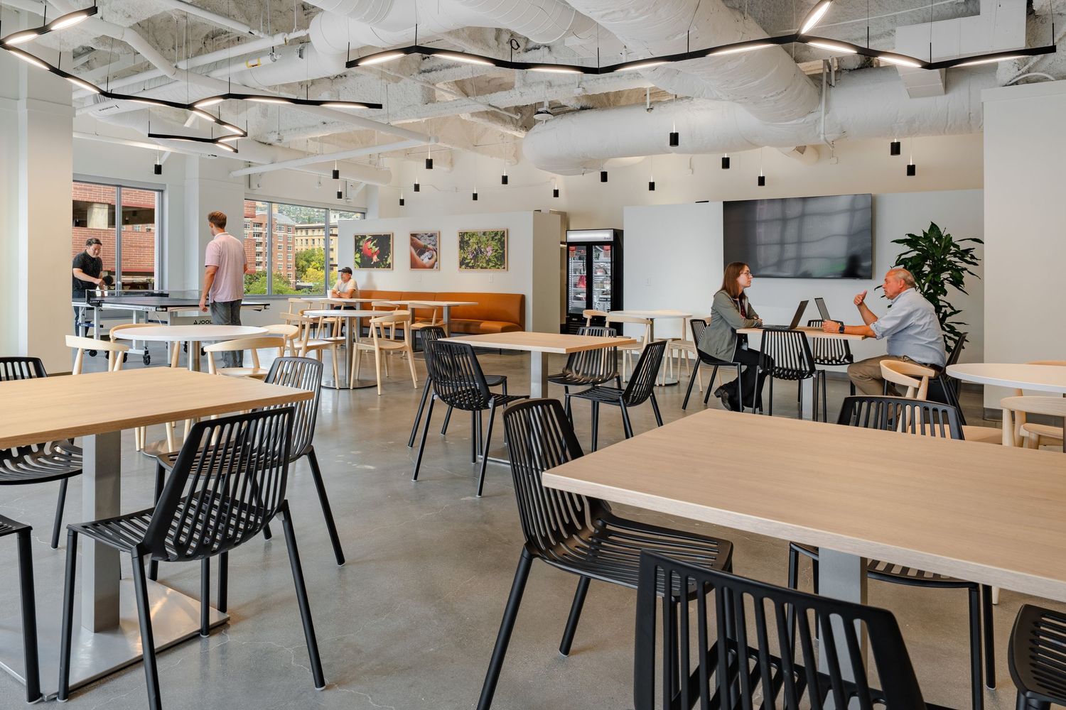 Modern office cafeteria with wooden tables, black chairs, exposed ceiling, and natural lighting through large windows.