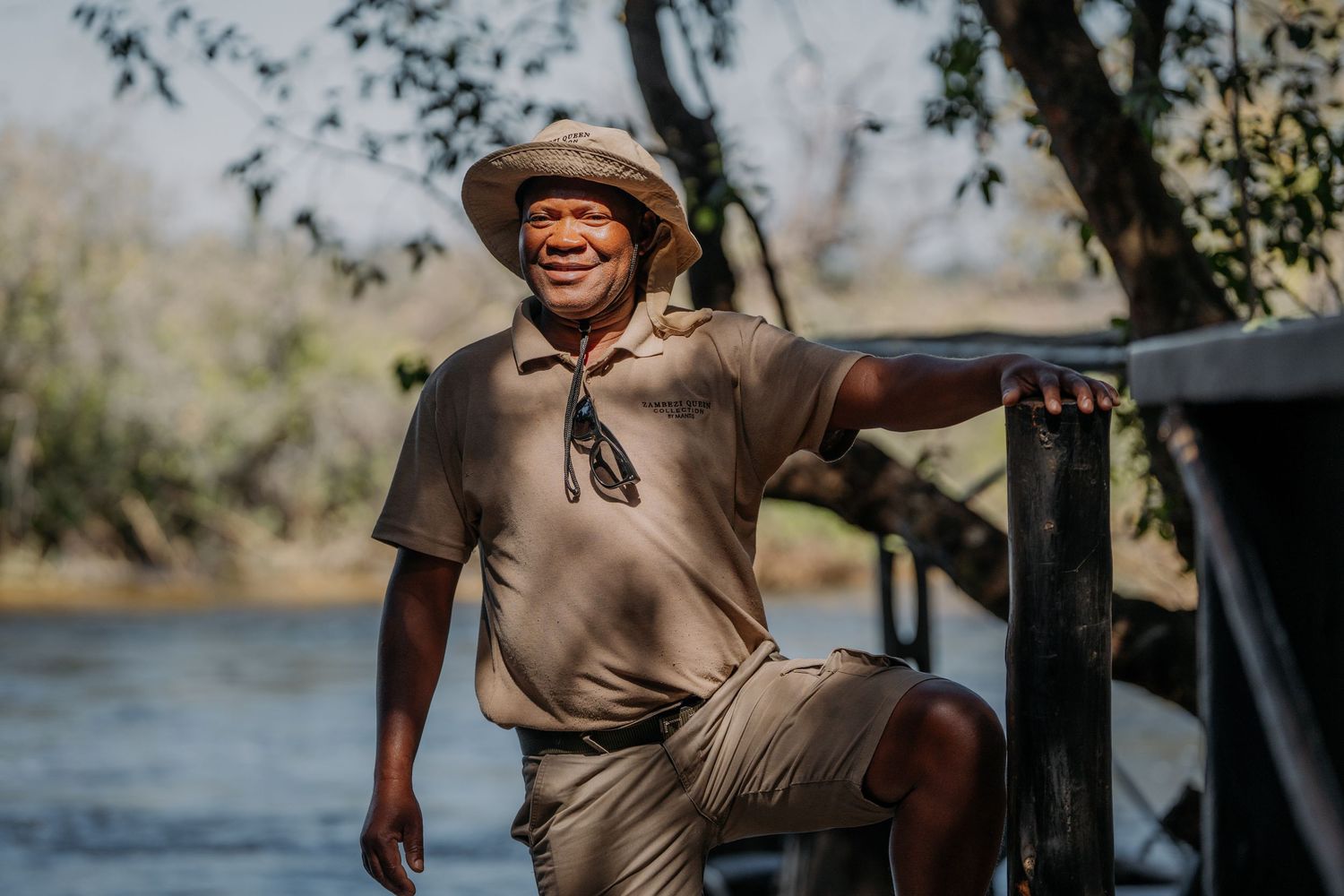 Smiling man in safari hat and khaki outfit leaning on railing by a river in a sunny African landscape.