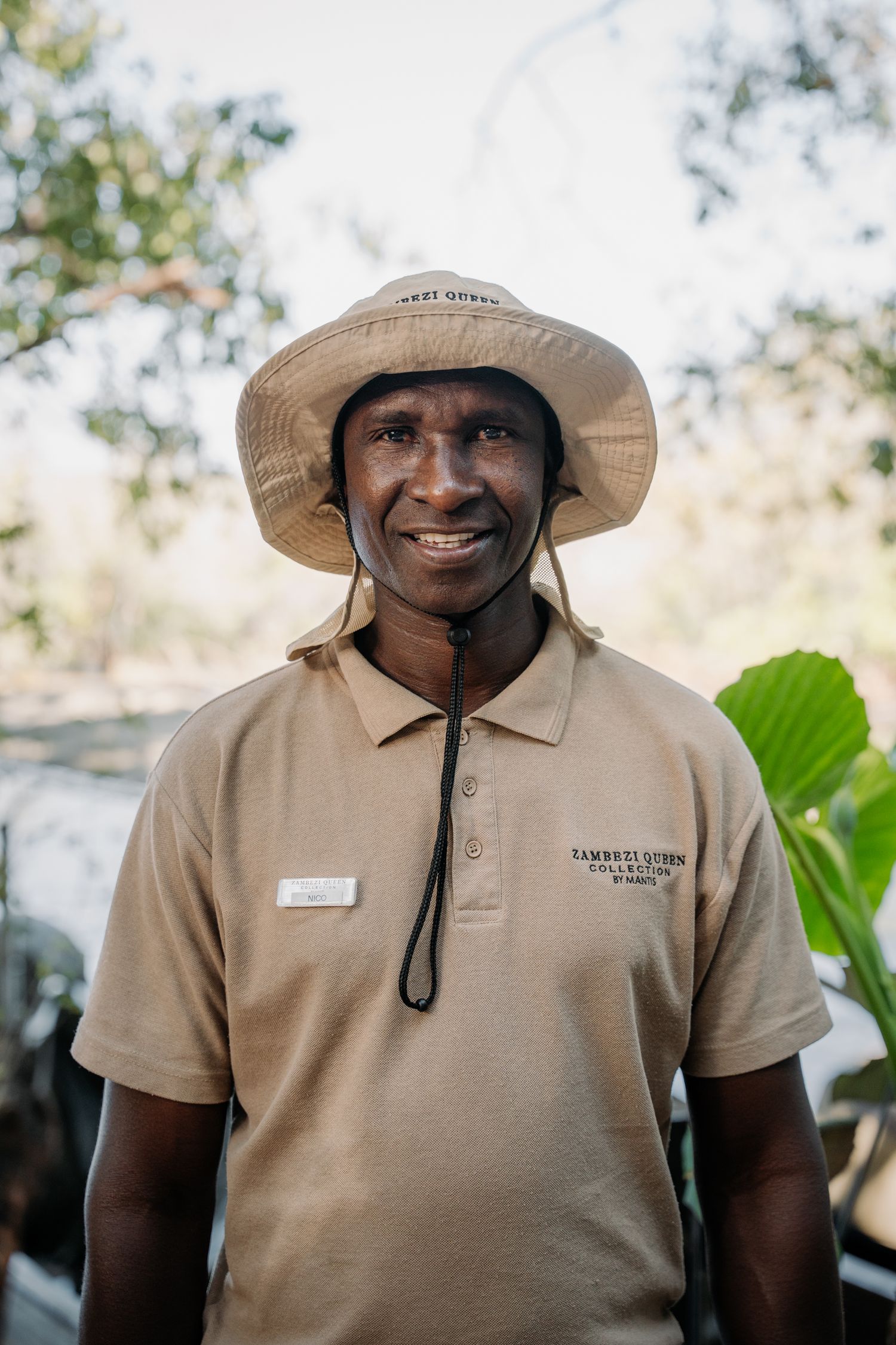 Smiling African safari guide wearing a tan uniform and bush hat with chin strap outdoors.