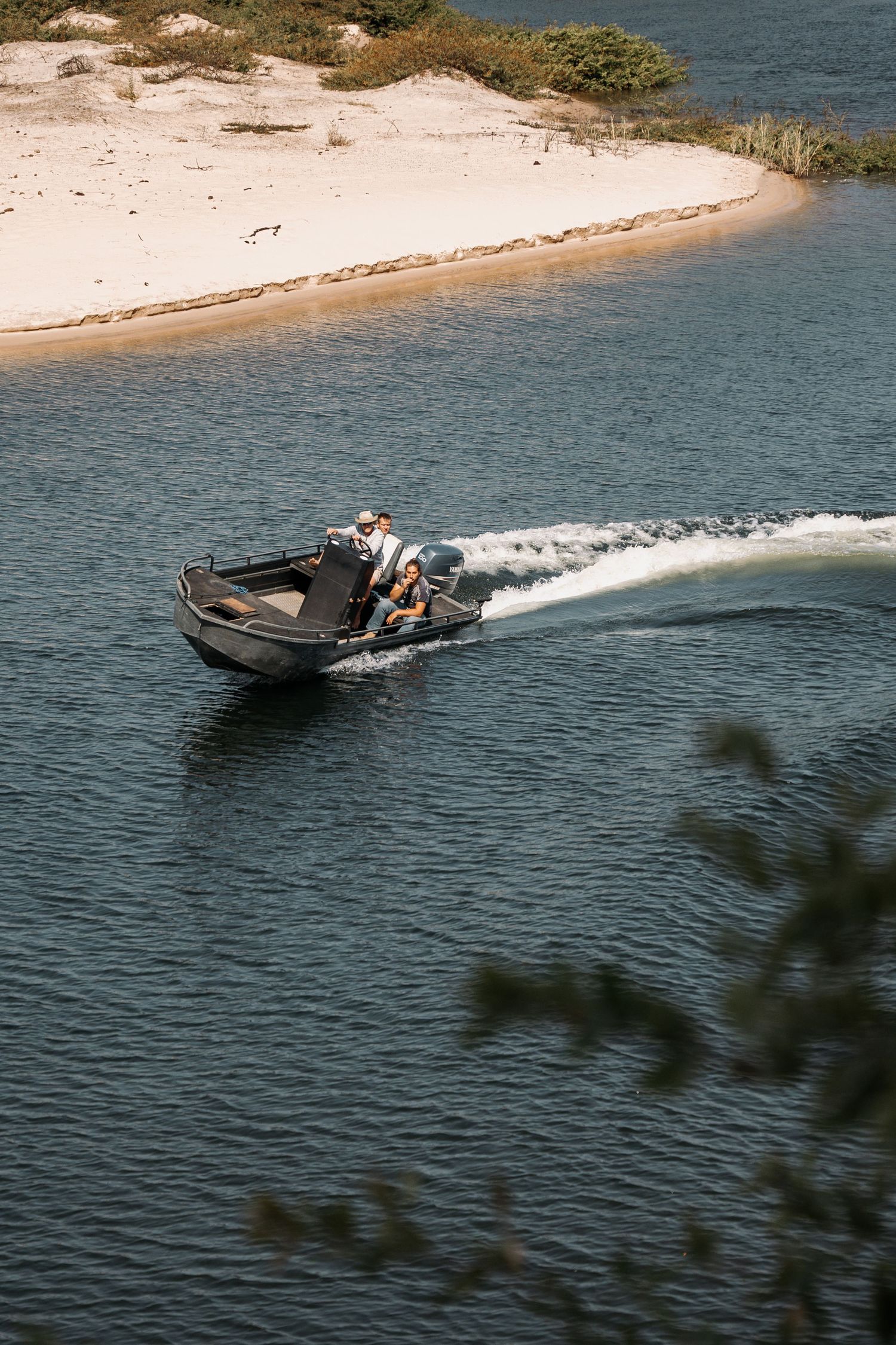 Speedboat racing across blue waters near a sandy shoreline creates white wake on a sunny day.