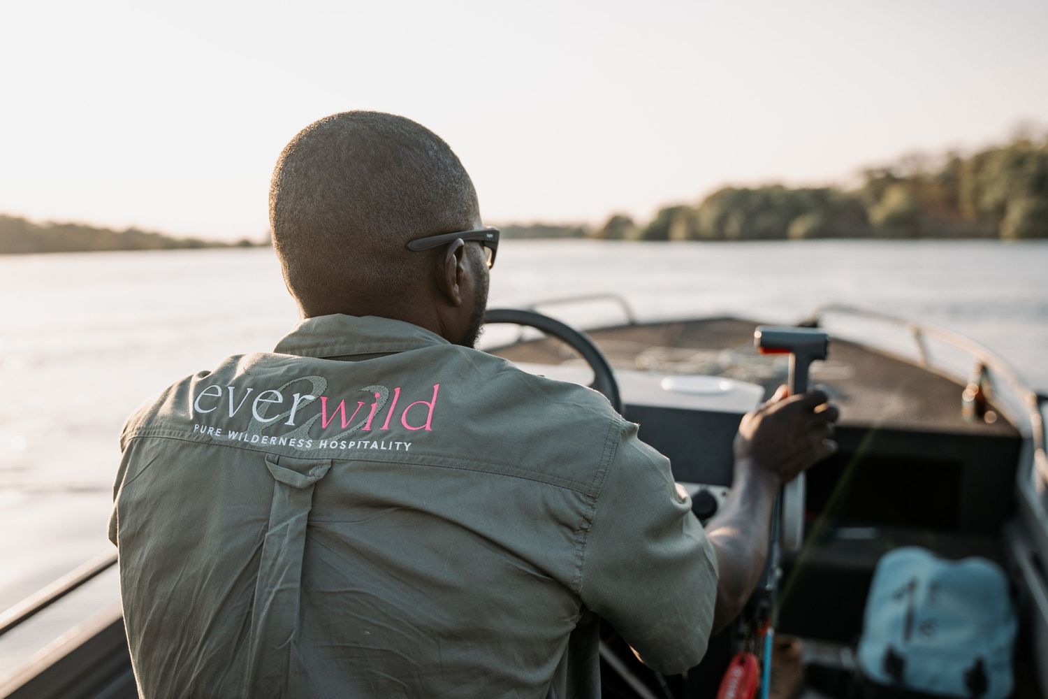 Person in glasses wearing a grey jacket steers a boat across calm waters at sunset.