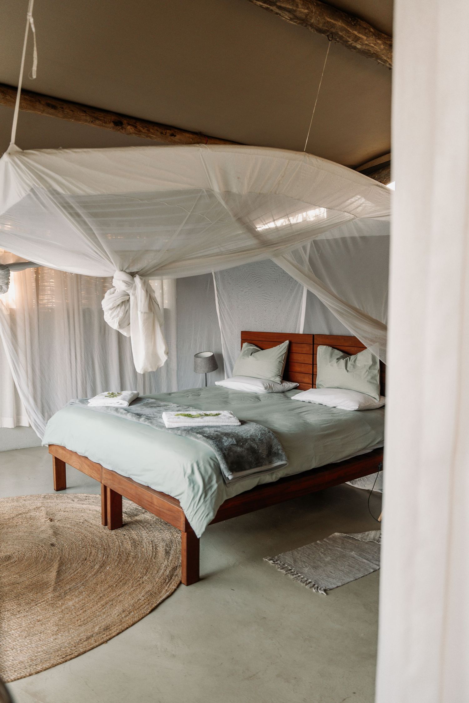 Rustic bedroom with white mosquito netting over wooden bed frame, light bedding and circular jute rug on concrete floor.
