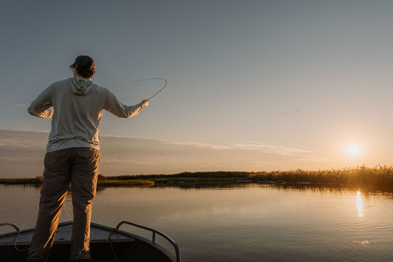 Man fly fishing from a boat at sunset on a calm lake with golden reflections on the water.