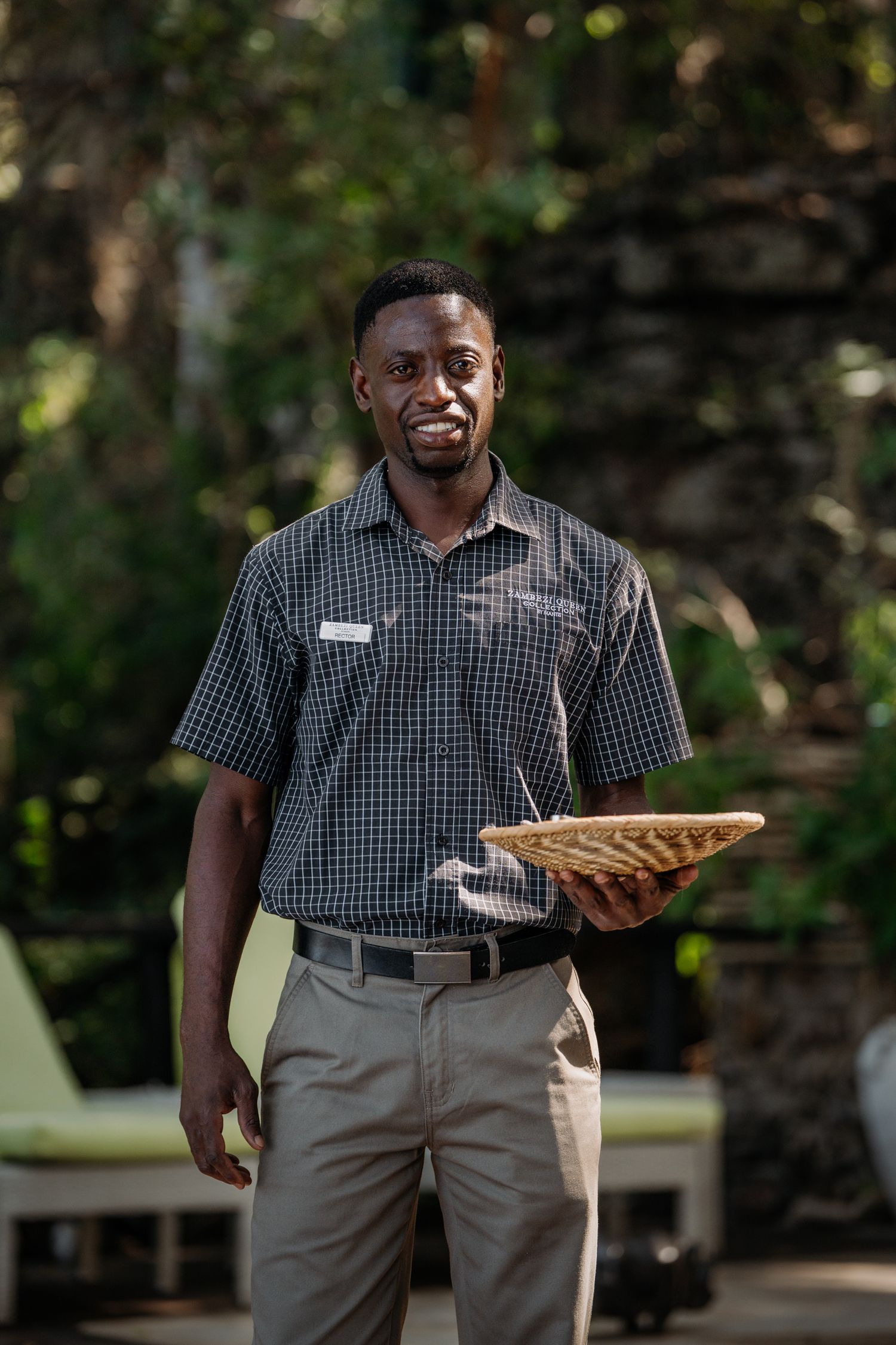 Smiling African lodge staff member in checkered uniform holding a woven basket tray outdoors.