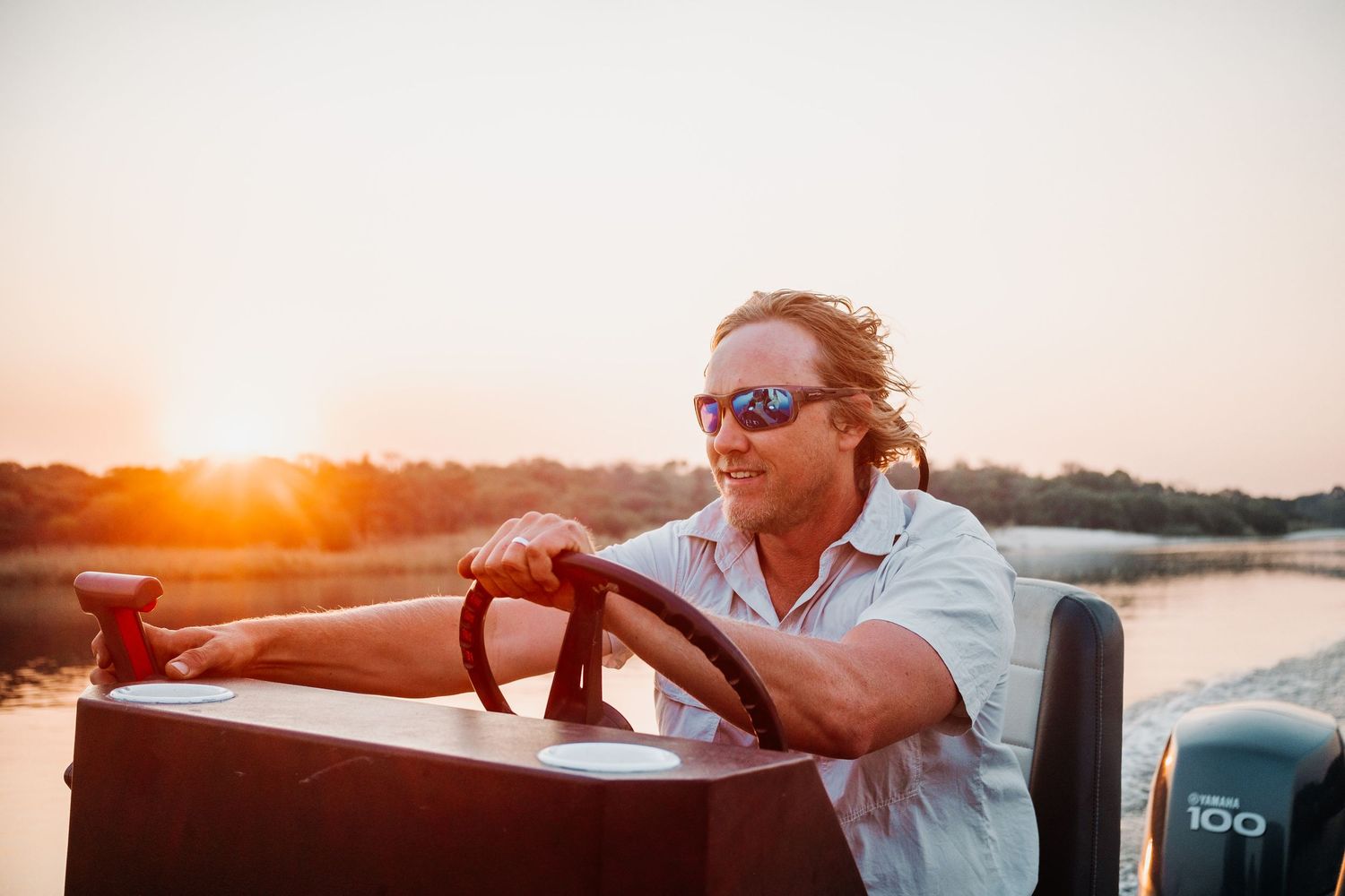 Person in sunglasses steering a boat during golden sunset over tranquil waters.