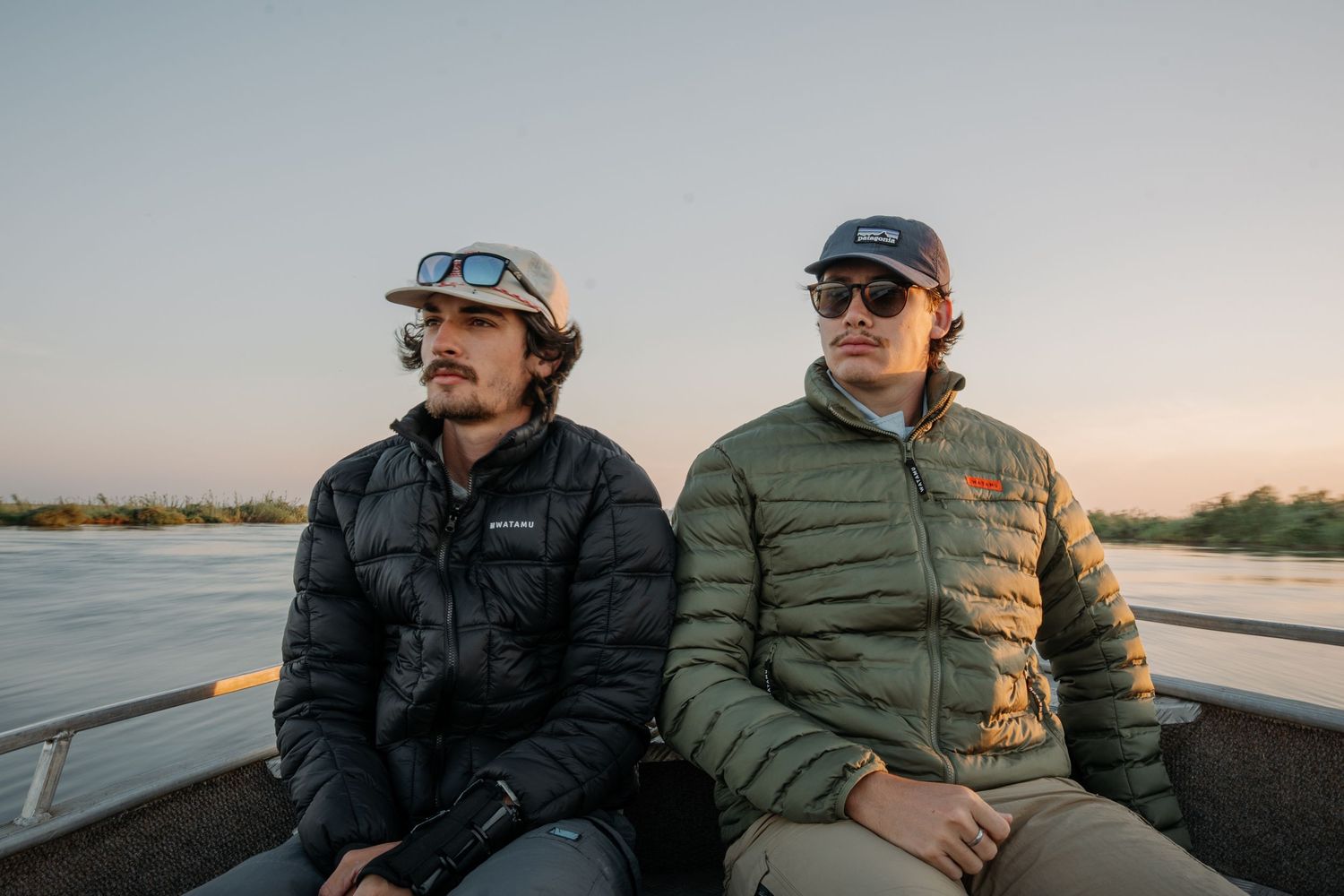 Two men in puffer jackets and caps sit on a boat at sunset near a calm river.