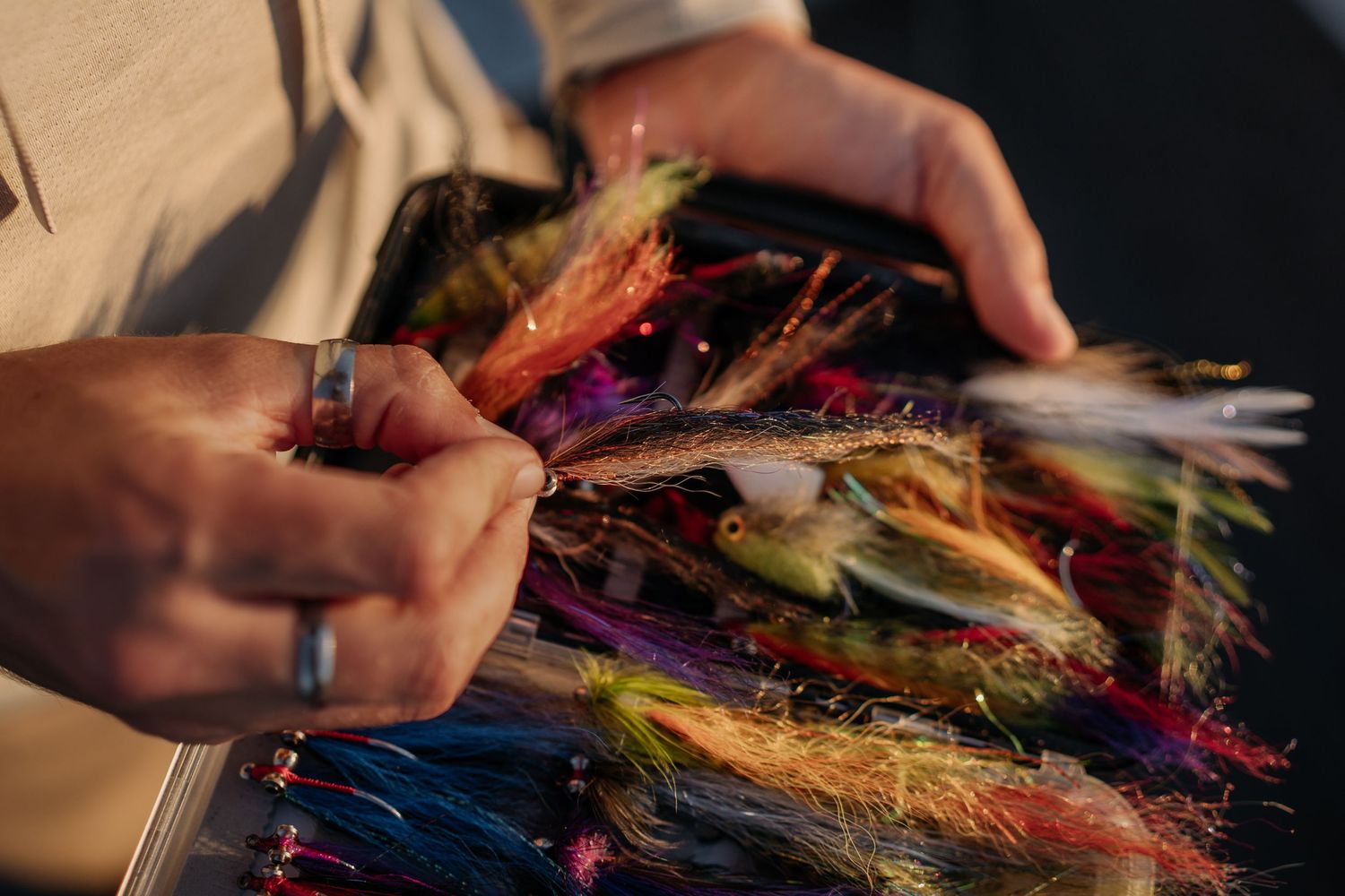 Hands sorting colorful fly fishing lures in a tackle box, featuring vibrant feathered flies for fly fishing.
