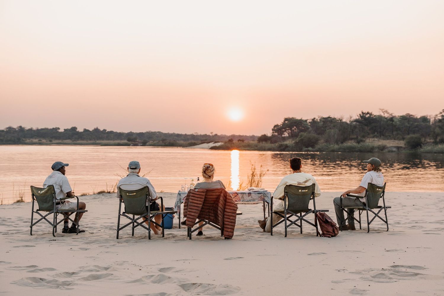 Group of people sitting in folding chairs watching a beautiful sunset over a river while having an outdoor gathering.