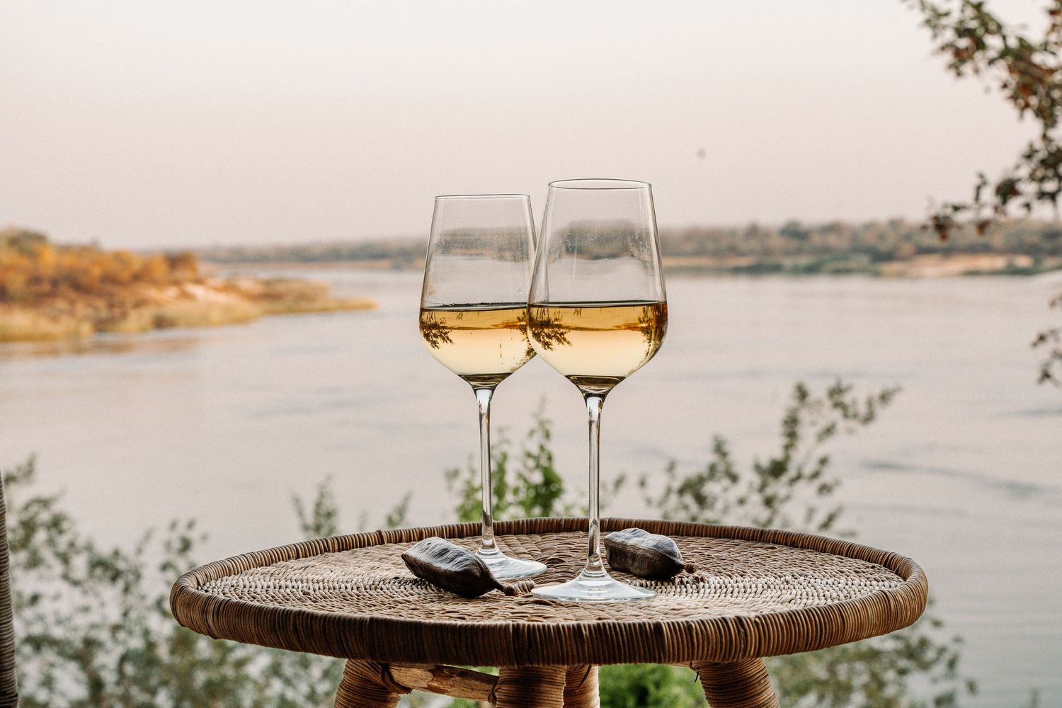 Two wine glasses filled with white wine sit on a wooden table overlooking a scenic waterfront view at sunset.