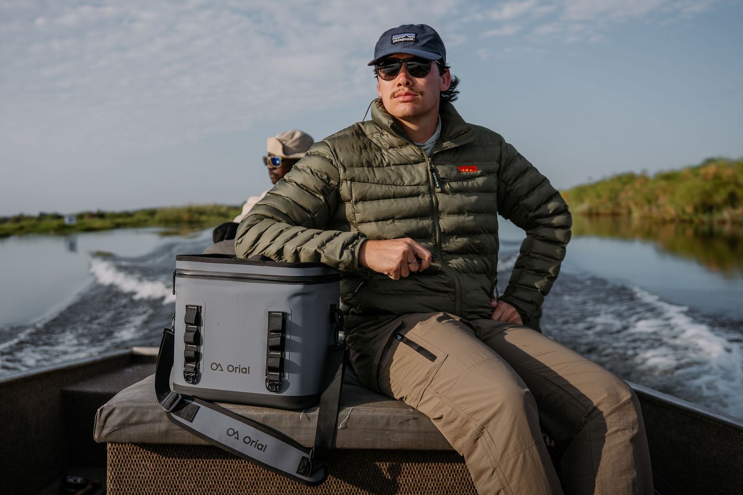 Man in olive jacket sits on a boat with a gray cooler bag, cruising through a marsh waterway at sunset.