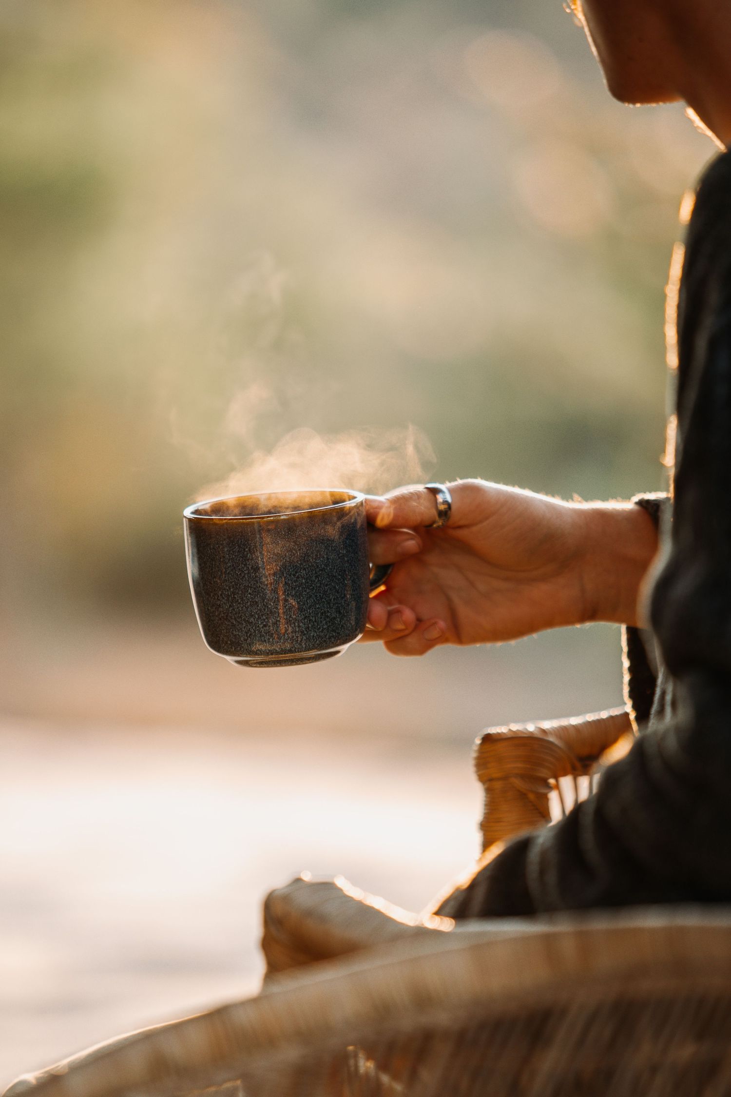 Person holding a steaming dark ceramic mug outdoors in warm golden light, enjoying a hot beverage in nature.