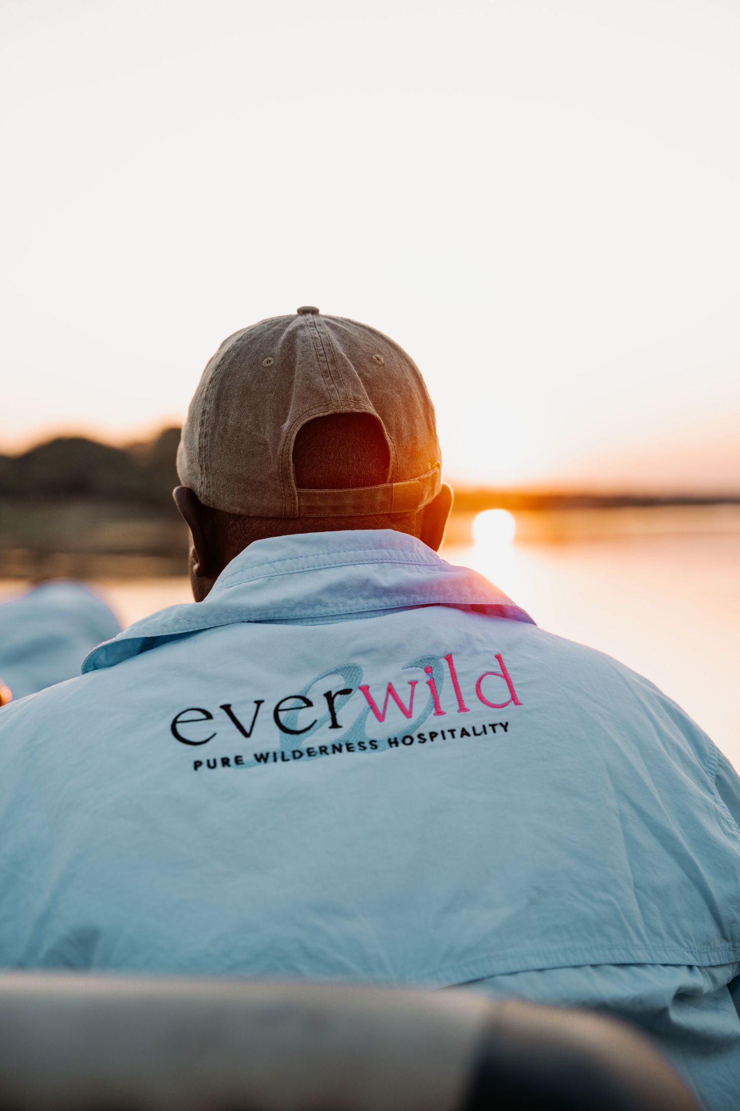 Person wearing an Everwild Pure Wilderness Hospitality shirt and cap watching a serene sunset over water.