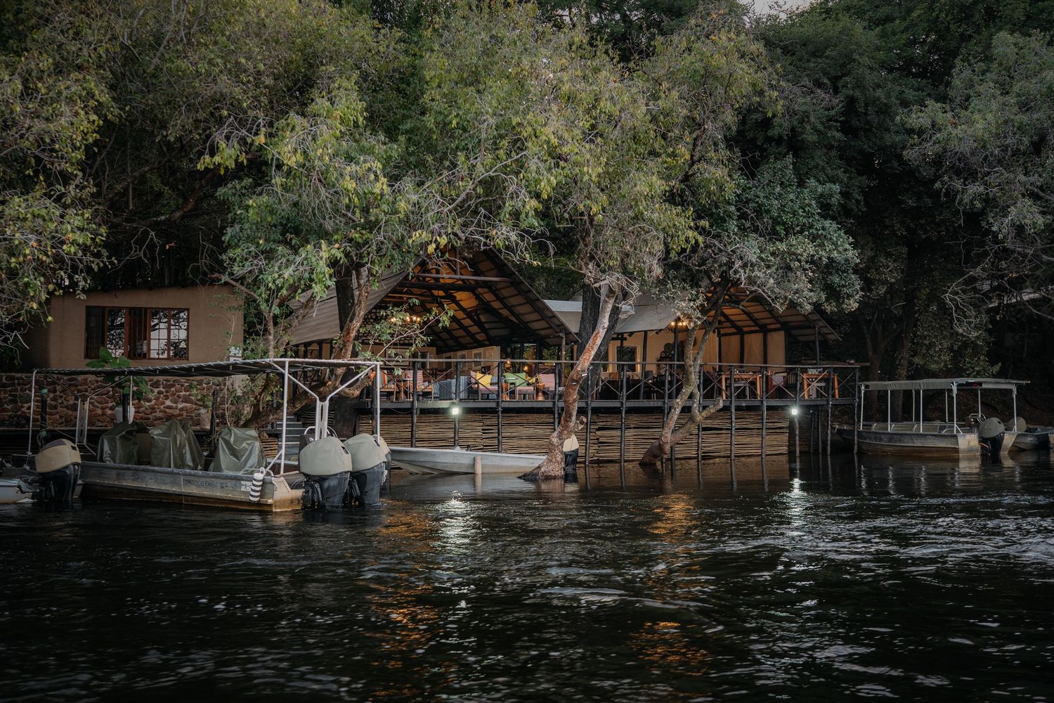 A riverside lodge with wooden dock, boats, and lush trees reflected in calm water at dusk.