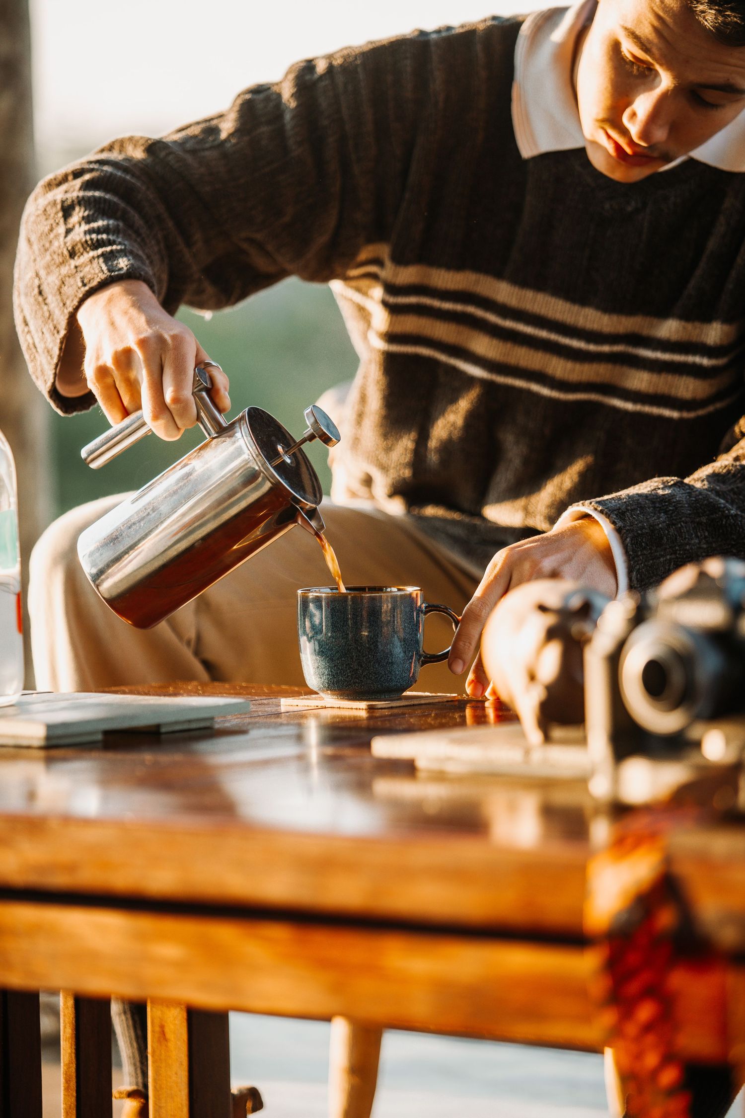 Someone wearing a striped sweater pours hot coffee from a French press into a blue ceramic mug on a wooden table.