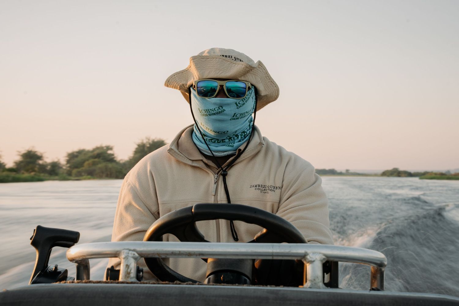 Man in hat, sunglasses, and face gaiter steering a boat on a calm river at dusk.