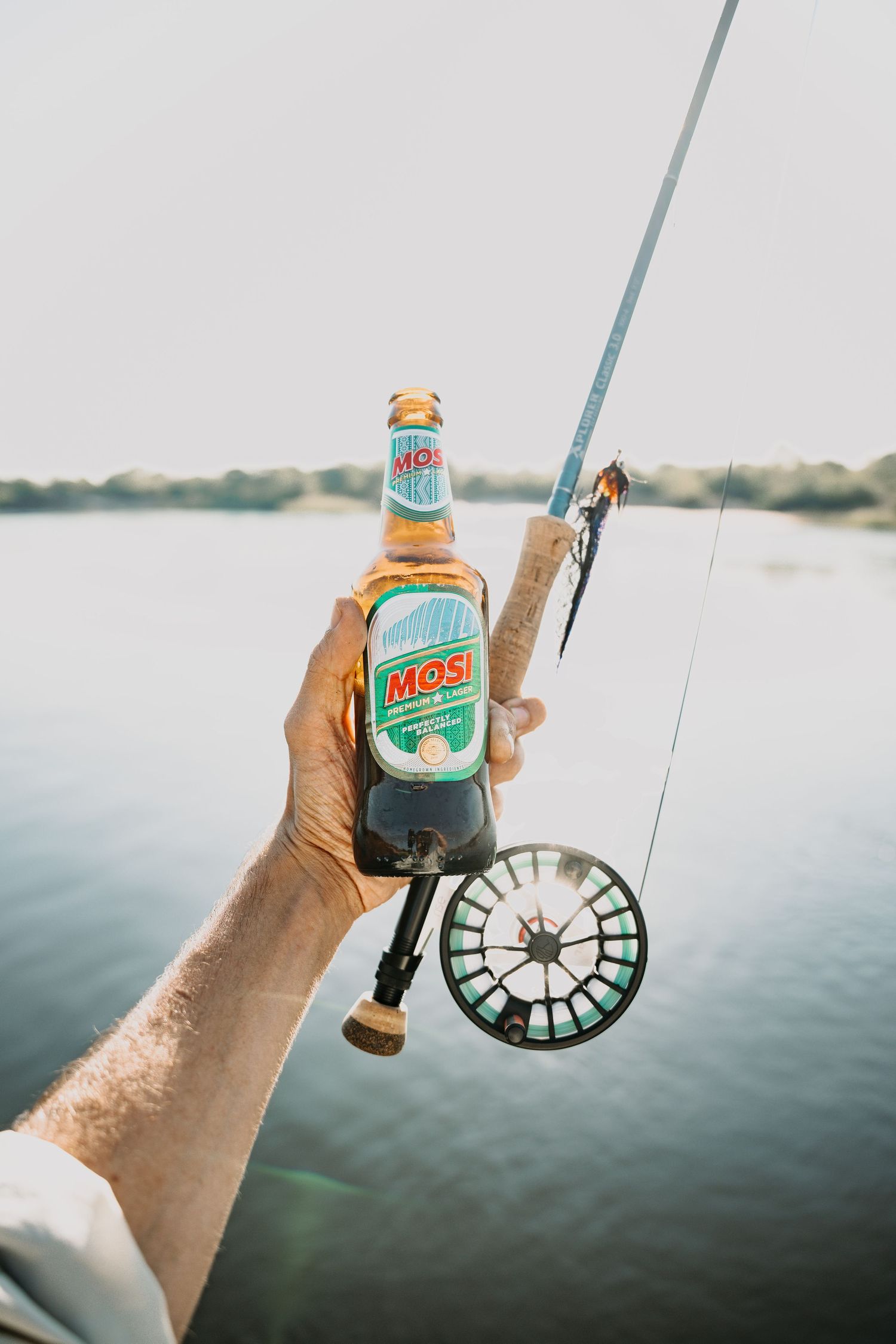 Hand holding a Narragansett beer can against a serene water backdrop with fishing rod and reel.