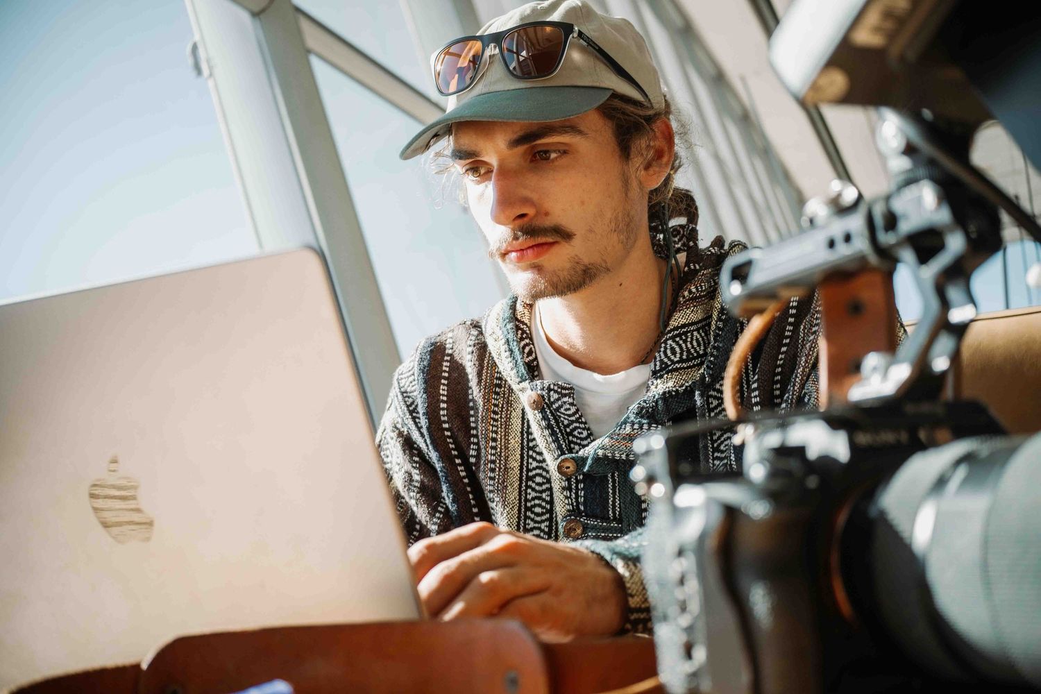 Someone in a hat and hoodie works on a MacBook laptop next to professional camera equipment.