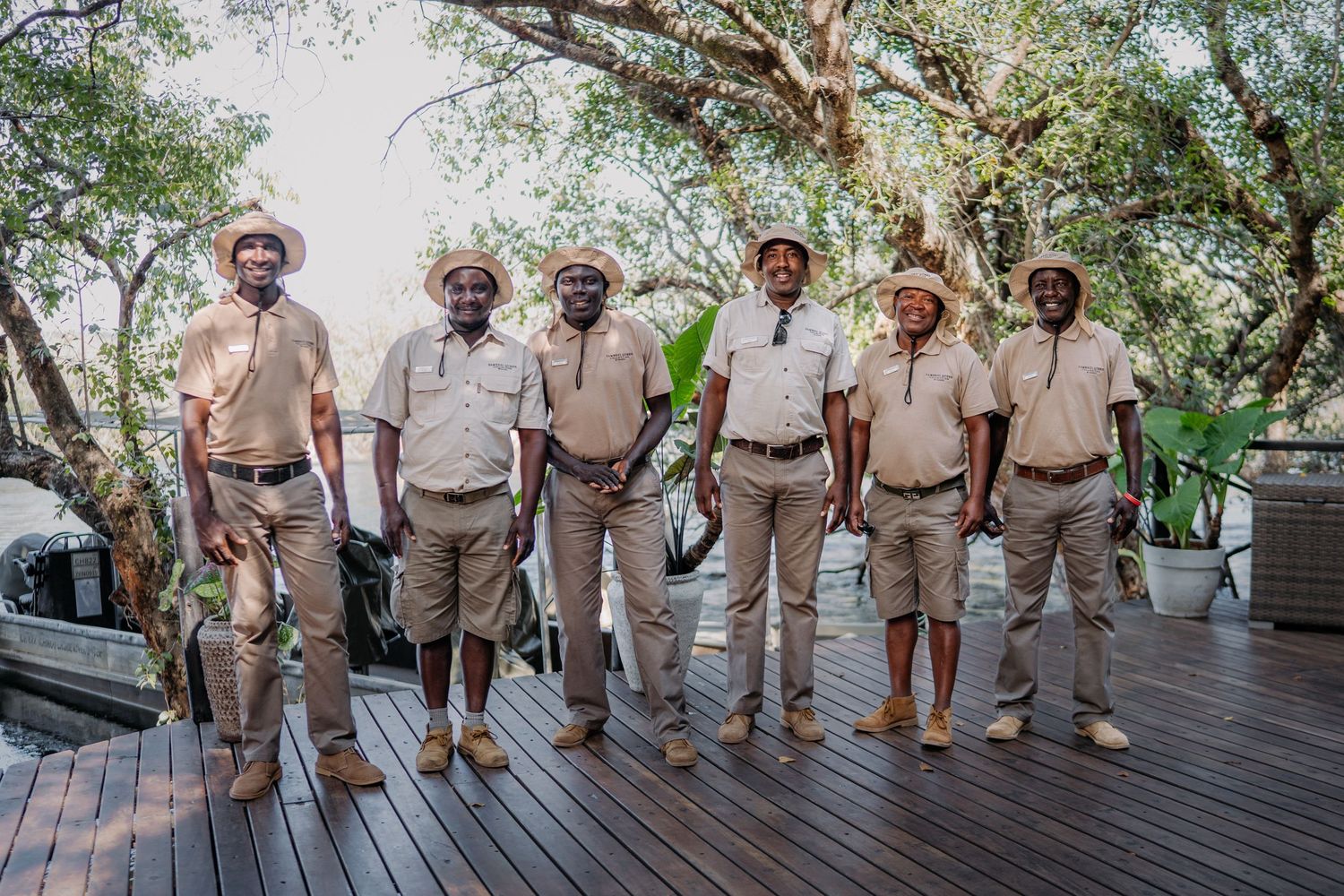 Six safari guides in matching khaki uniforms and straw hats posing together on a wooden deck surrounded by trees.
