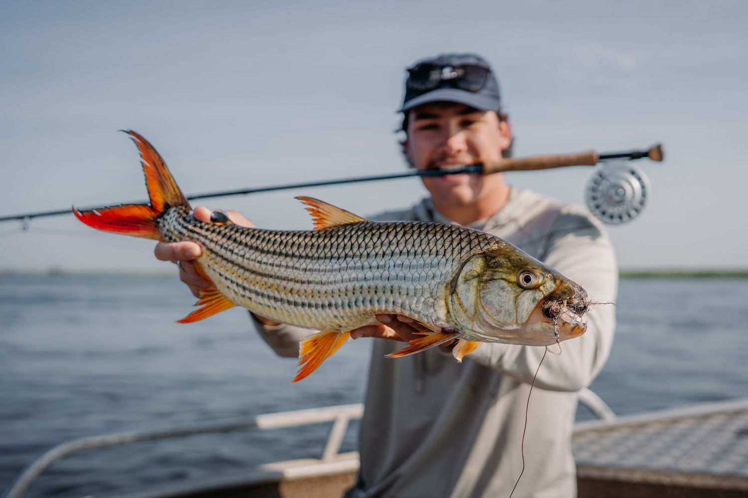 Smiling angler holds a vibrant tarpon fish on a boat, with a fly fishing rod and open water in the background.