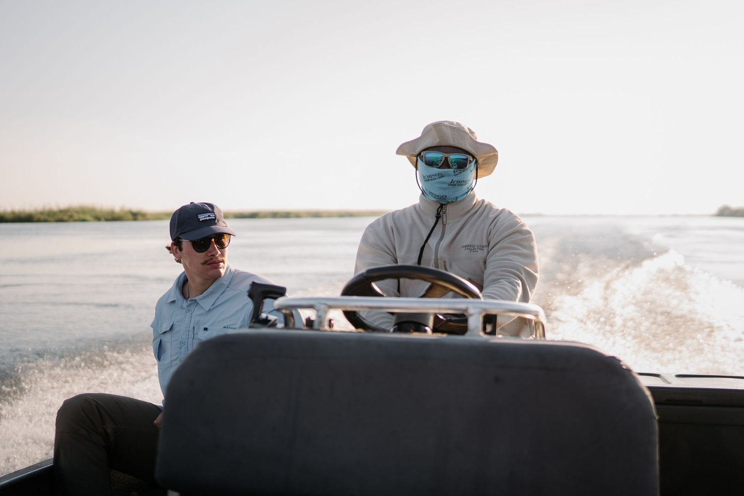 Two men wearing masks and hats ride a speedboat across open water on a sunny day.