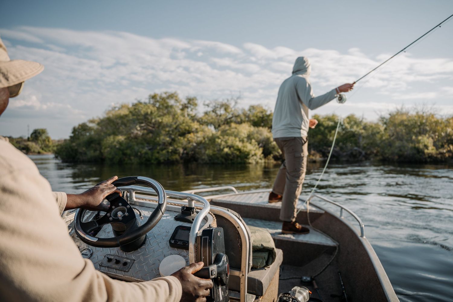 Two men fishing from a motorboat on a calm river surrounded by lush green trees under a partly cloudy sky.