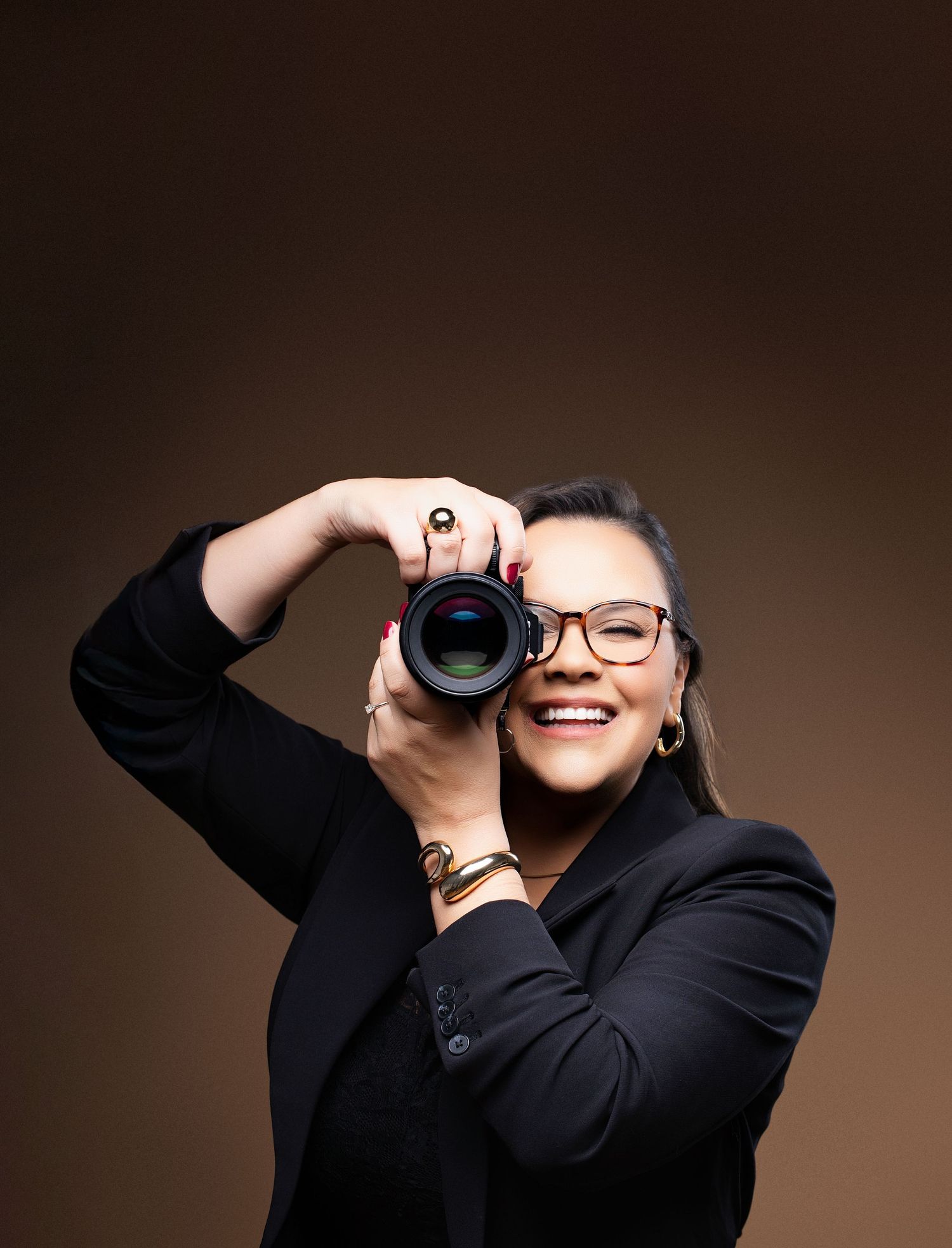 Professional photographer wearing black suit and glasses holds camera up to eye while taking photos against dark background.