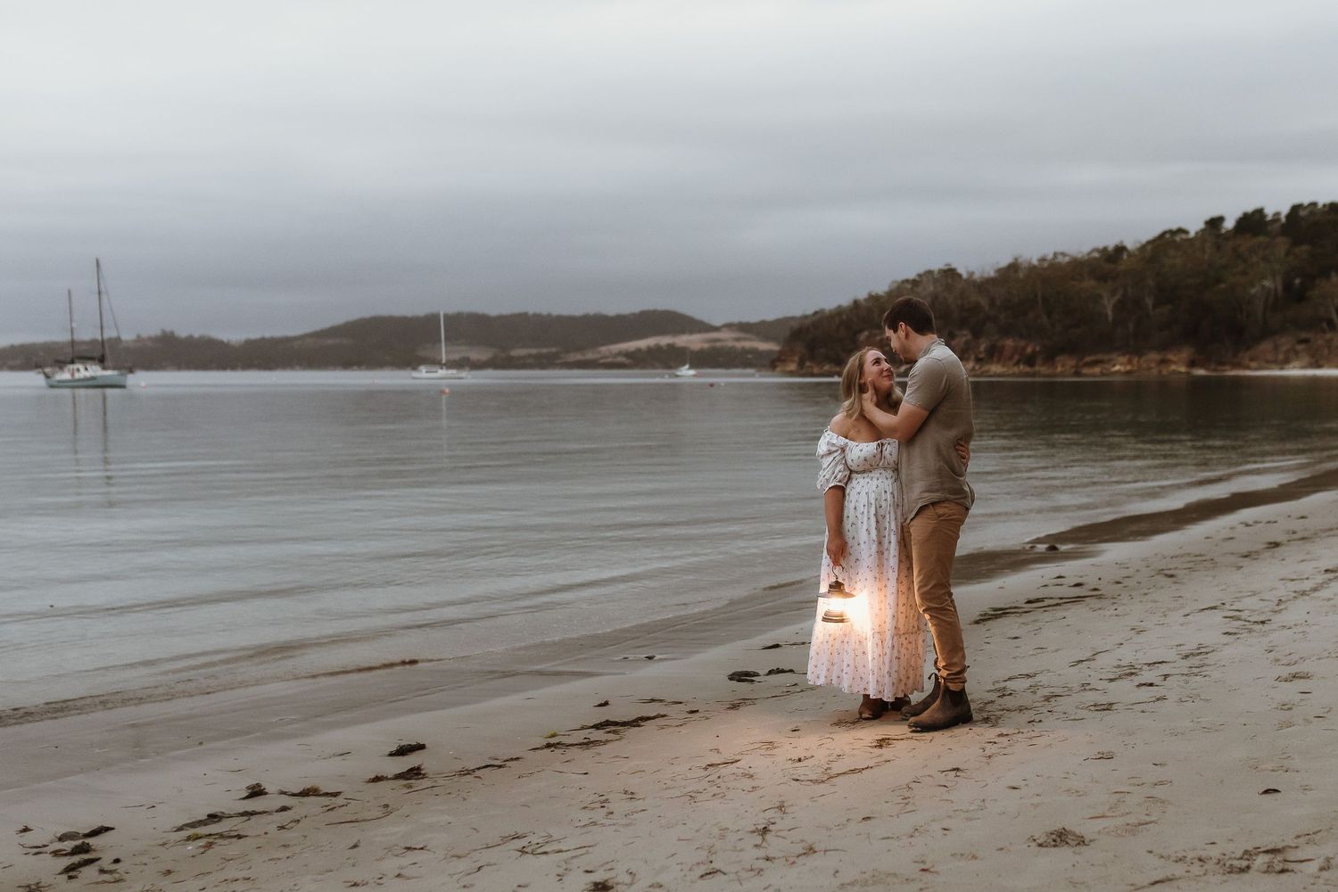 Couple embracing on a sandy beach at dusk, holding a lantern with sailboats and hills in the background.