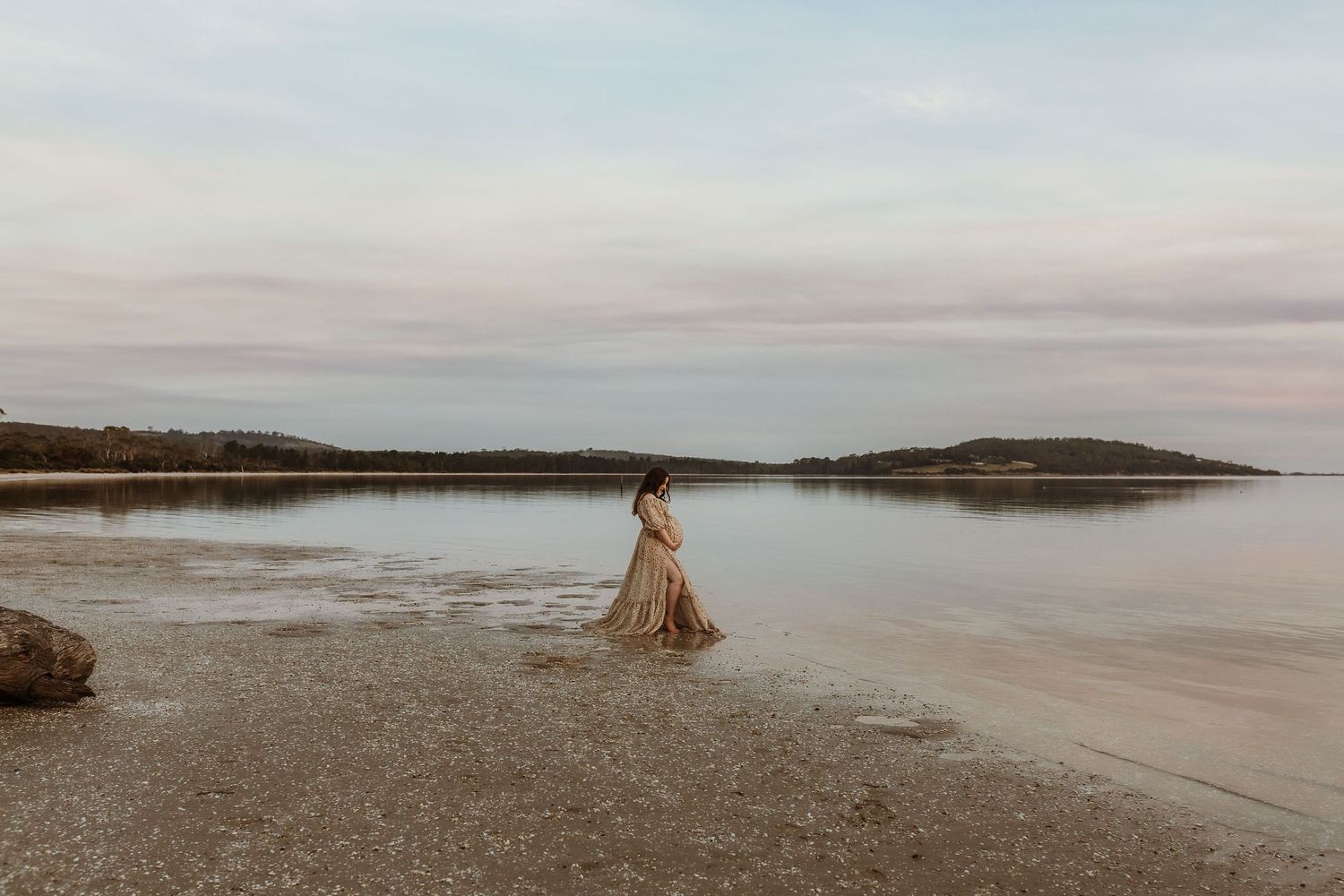 A woman in a flowing golden gown stands in shallow coastal waters at dusk with a serene landscape behind her.