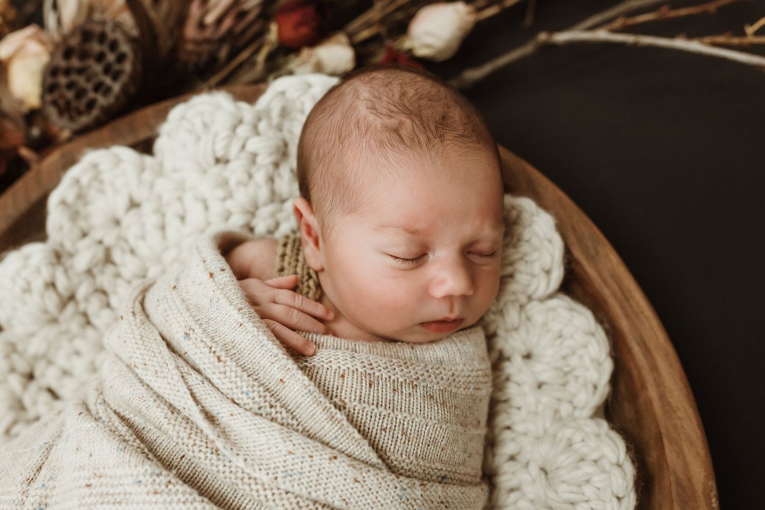 Sleeping newborn baby swaddled in cream knit wrap, nestled in wooden bowl with chunky crochet blanket.