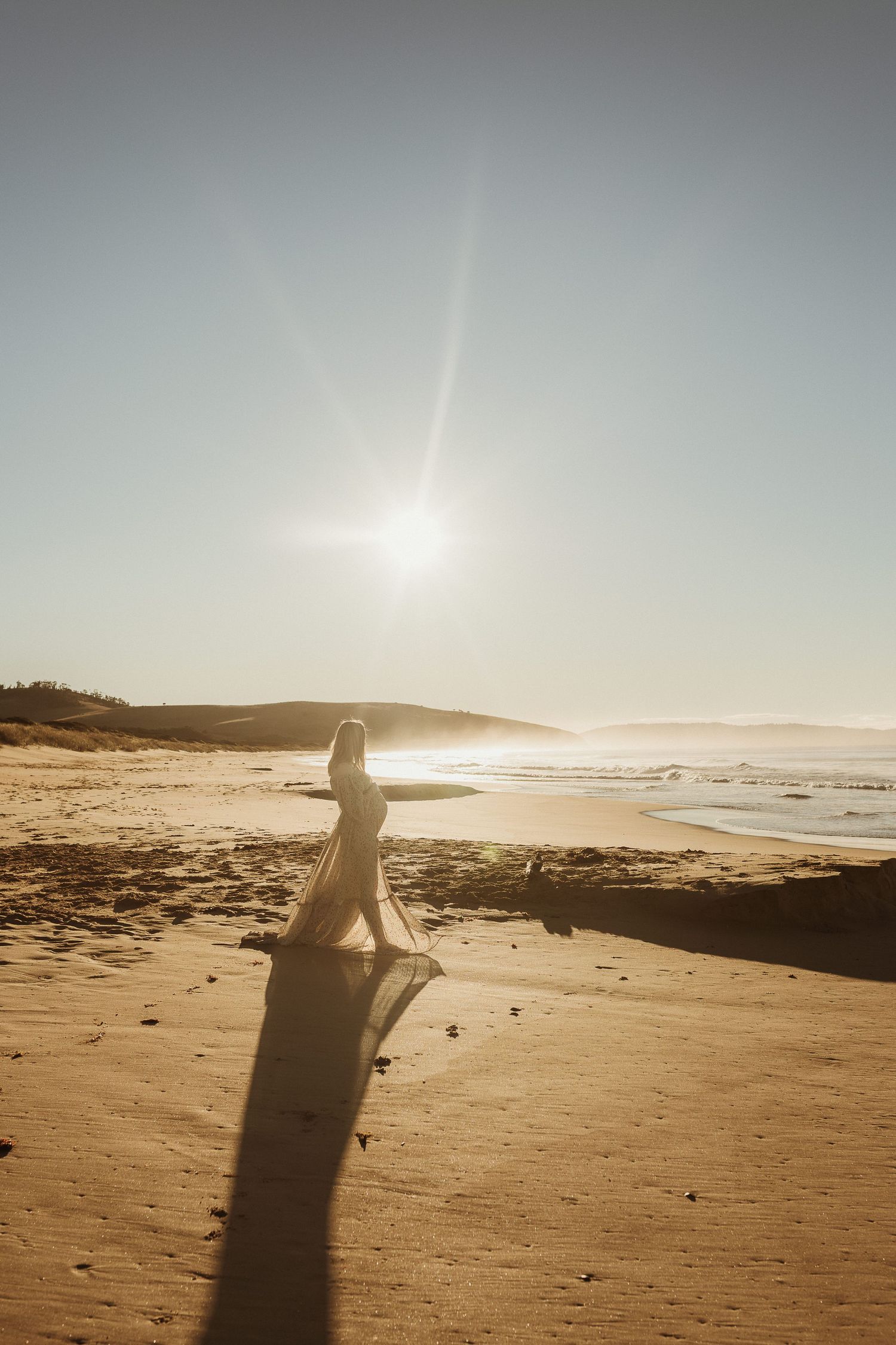 A person's silhouette casts a long shadow across a sandy beach at sunset with ocean waves in the background.
